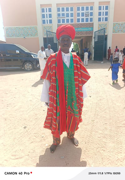 A full shot captures a man standing barefoot on a sandy expanse. He wears a red turban, a white shirt, a vibrant green embroidered vest, and a red and yellow striped outer garment. Behind him, a pale building with blue and white windows and decorative art stands. A black van is parked to the left, with people milling around the building's entrance and in the background. The location is Langtang, Nigeria.