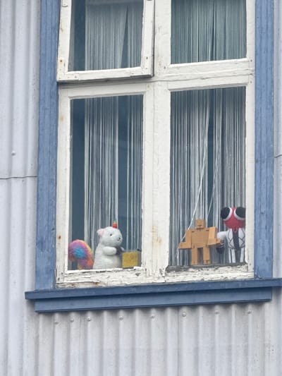 The image displays a detailed exterior view of a building in Reykjavik, Iceland, focusing on a multi-paned window. The building's facade is constructed from light gray corrugated metal siding. A faded blue trim outlines the window and forms a sill beneath it. The window frame is painted white, exhibiting signs of age with peeling paint and minor rust stains, particularly on the left vertical section. Thin, white, string-like curtains or blinds are visible behind all window panes.
In the bottom left window pane, a white plush toy, resembling a unicorn with a small, rainbow-striped horn, stands. A distinct rainbow-colored fluffy tail is visible to its left. The toy appears to be positioned next to a small, yellow, rectangular object. In the bottom right window pane, a light brown, blocky, wooden figure, possibly a stylized human or robot, hangs suspended by thin white strings. This figure has a visible cross-shaped indentation on its chest. To its right, partially obscured by the window frame, is a plush character identified as "Red" from the "Rainbow Friends" franchise, characterized by a spherical red head with large black and white eyes, and a white body with black vertical stripes resembling a tuxedo. This "Red" character is shown holding the left arm of the wooden figure.