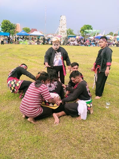 The scene takes place on a grassy field in Karangmalang, Indonesia, at coordinates -7.47 latitude and 111.064 longitude. Seven men are present, with five men in the center of the frame. There is a single empty glass cup on the ground near the right edge of the frame. The man at the center of the group is sitting on the ground, with his legs stretched out in front of him. His body is being held by three other men, one of whom is touching his chest with both hands. The man at the right of the frame is wearing black pants and a black shirt with a red sash and is standing. He is looking towards the center of the frame. There are tents and other structures in the background. The ground is covered in grass. The sky is overcast with clouds.