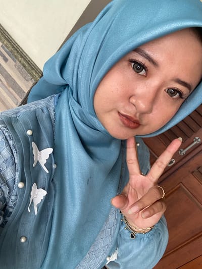 A selfie-style image depicts a young to middle-aged adult female positioned in the foreground, facing the camera with a slight head tilt and direct gaze. She has light brown skin, dark eyes with enhanced eyelashes, and is wearing brownish-orange lipstick. Her head is covered by a light blue, slightly shimmery hijab. She is dressed in a light blue garment, possibly a top or dress, featuring a textured or sheer overlay on the upper torso. This overlay is adorned with multiple white pearl-like beads and two white, fabric butterfly appliqués, each detailed with smaller white beads. Her right hand is raised to chin level, forming a "peace" or "V" sign with the index and middle fingers extended. On her right ring finger, she wears a gold-toned ring with small clear stones. Multiple gold-toned bracelets, including one with black heart charms, are visible on her right wrist. The background is partially visible and out of focus, showing a light-colored, possibly tiled or textured wall section on the left and a dark brown wooden surface, consistent with a door or paneling with metal hardware, on the right. The scene is located in Ketegan, Indonesia.