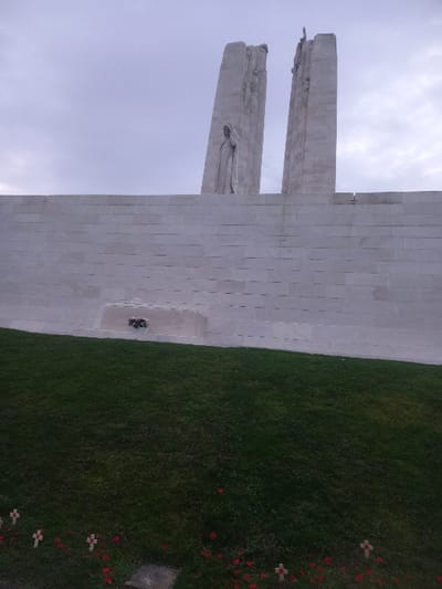 The image depicts the Vimy Memorial. Two imposing, light-colored stone pillars rise towards a cloudy sky. Carved figures are visible on the pillars. A long, light-colored stone wall extends to the right of the pillars. At the base of the wall is a recessed area with a wreath. Green grass covers the ground. Small white crosses adorned with red poppies are scattered on the right, near the grass's edge.