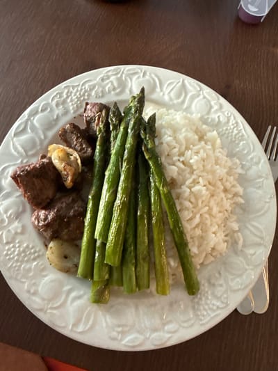 The image presents an overhead, close-up view of a prepared meal served on a white, round plate with ornate, embossed decorative patterns, possibly depicting grapes and vines. The plate rests on a dark brown surface, which appears to be a wooden table.
The meal consists of three main components: a substantial serving of white, fluffy rice positioned towards the top of the plate, a neatly arranged stack of vibrant green, cooked asparagus spears lying horizontally across the middle, and several browned cubes of meat, likely beef, situated at the bottom of the plate. Alongside the meat, there are a few lighter-colored, softened pieces that resemble sautéed garlic or onion.
A silver fork is partially visible in the upper right corner of the frame, indicating the meal is ready to be eaten. In the blurred upper-left background, a small, purple and white object can be seen, possibly part of a container or bottle. The scene depicts a domestic setting, likely a dining area, and suggests a home-cooked meal. The location context is Marche-en-Famenne, Belgium. No text is visible in the image.