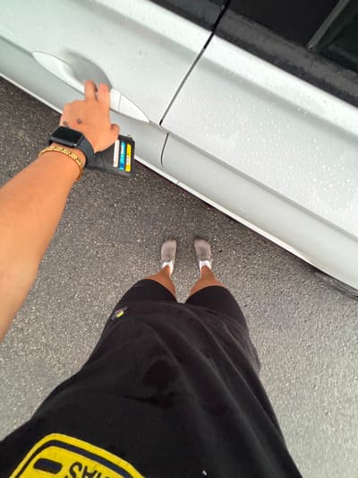 A person in black shorts, a black t-shirt, and grey shoes stands beside a white car. They're holding a black wallet near the car door handle. The person is wearing a gold bracelet and a smartwatch on their left wrist. Raindrops are visible on the car's surface and the asphalt. The t-shirt has a yellow patch visible. The photo appears to be taken from an overhead perspective.
