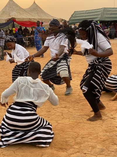 Three people are dancing outdoors. They all wear white shirts with black and white striped skirts. The dancer at the top of the image has black leggings under their skirt and is barefoot. The dancer in the middle has gray sneakers. All dancers are facing the ground and appear to be in motion. A crowd watches in the background underneath a tent.