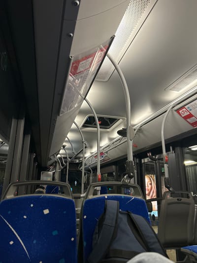 The inside of a bus features blue patterned seats, a dark gray aisle floor, and overhead handrails. Advertisements are visible on the windows. The bus ceiling is light gray with ventilation grates. A transparent panel displays transit information with a red CTS logo. A backpack rests on a seat. The view suggests the bus is in motion at night.