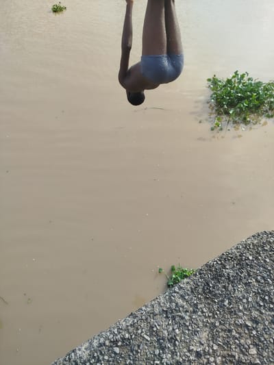 A person in grey swimming trunks is captured mid-air, upside down, about to enter murky brown water from an elevated concrete structure. The person's arms are extended upwards, and their legs are bent at the knees. Green aquatic plants are visible floating on the surface of the water. The scene appears to be in Yenegoa, Nigeria, likely a natural waterway or canal edge where such recreational activity is possible.