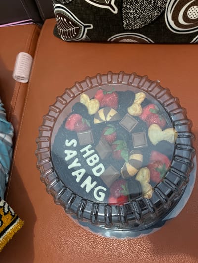 A circular chocolate cake with white icing letters spelling "HBD SAYANG" is displayed in a clear plastic container. The cake is decorated with strawberries, chocolate squares, cookies, and possibly black cherries. The cake is placed on a brown leather surface, likely a couch. In the background, a patterned throw pillow and a pink folded paper are visible. To the left of the cake, a small white cylindrical object, possibly a hair roller, rests on the brown surface. A portion of a blue and white checkered fabric is also visible. The scene is an indoor setting in Jambearum, Indonesia.