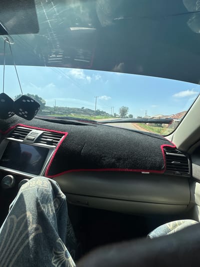 The image captures the interior of a car, looking out through the windshield. In the foreground, a black dashboard cover with a red trim is visible, along with two fuzzy black dice hanging from the rearview mirror. The passenger's lap, covered in patterned denim, is also in the frame. Through the windshield, a road is seen stretching into the distance, flanked by sparse vegetation and a few buildings with corrugated roofs. Utility poles and power lines are present along the roadside. The sky is blue with scattered white clouds. The scene appears to be in a rural or semi-urban area, consistent with the description of Agingi Zion, Nigeria. The image is taken from the passenger seat.