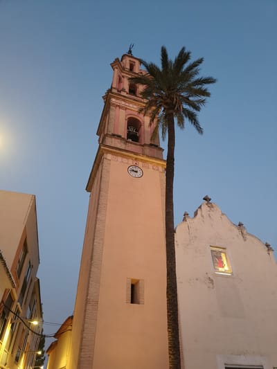 The image shows a view of the Church of Sant Bartomeu i Sant Roc in Benirredrà, Spain, against a twilight sky. The church features a prominent bell tower with a clock and a pink facade. A tall palm tree stands in front of the tower, partially obscuring it. To the left of the bell tower, another building with a light-colored facade and windows is visible. The right side of the image shows the facade of the church with a stained-glass window depicting a religious figure. Streetlights are visible on the left side of the frame, illuminating the facades of buildings in what appears to be a narrow street. The overall scene suggests a calm evening in the town.