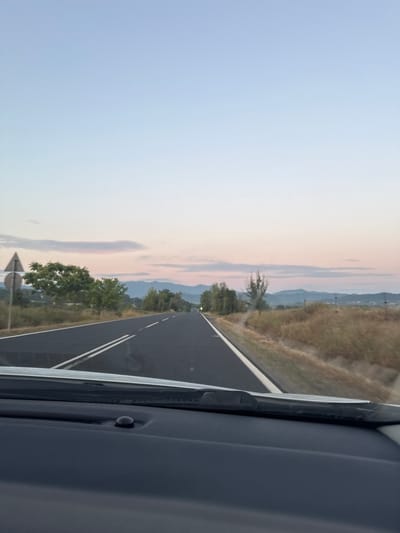The image shows a road in Varben, Bulgaria, at dusk. The road is paved with asphalt and has two white lines marking the lanes. There are trees and dry grass on the side of the road. In the distance, there are mountains. The sky is clear with a few clouds. The image is taken from inside a car, looking out the windshield.