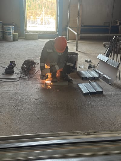 The image depicts an indoor construction or workshop setting in Chulman, Russia. A worker wearing a red hard hat, a reflective vest over a grey uniform, and work boots is crouched on a concrete floor. The worker is actively using an angle grinder, generating sparks as they cut through metal framing material. Nearby, a bucket, a power tool with battery packs, and electrical cords are visible. Several stacks of U-shaped metal profiles are arranged on the floor. In the background, a large window offers a view of a tree-lined outdoor area with sunlight. Scaffolding and building materials are also present in the background, suggesting ongoing construction or renovation.