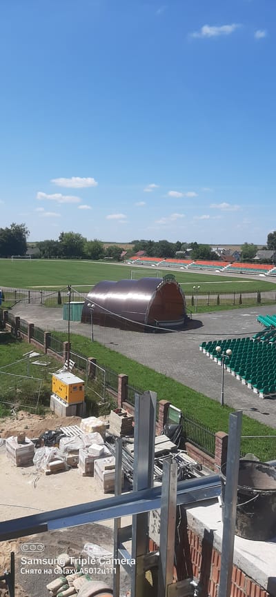 This is a medium-angle, vertical, outdoor shot of a sports stadium and its surrounding area on a clear, sunny day. In the foreground, a construction site is visible with piles of bricks, pallets of bagged material, and metal framing structures. Further back, a yellow generator sits on a concrete base.

Beyond the construction area, a black metal fence separates the foreground from a paved walkway and a large, brown, arched structure, possibly a stage or shelter. To the right of this structure, rows of empty green stadium seats are arranged in a curved pattern.

In the middle ground, a well-maintained green sports field with white markings is visible, bordered by trees and some houses in the distance. Stadium seating in orange and red is also visible in the middle ground, along the curve of the field. The sky is a clear blue with a few white clouds.

The image likely depicts a sports facility in Vyalikaya Byerastavitsa, Belarus, undergoing renovation or expansion.