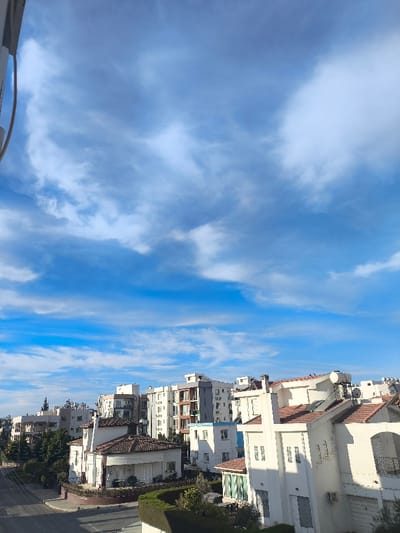 This image presents an elevated, daytime view of a residential area within Nicosia, Cyprus. The upper two-thirds of the frame consist of a bright blue sky with scattered cirrus and stratocumulus clouds. The lower third features urban infrastructure.
On the immediate right, a three-story, white-stucco building with a reddish-brown tiled roof is prominent. Its roof supports one satellite dish and two visible solar water heating tanks. The building displays several windows, some with closed white shutters, and an arched opening on an upper floor. A manicured dark green hedge runs across the lower foreground, partially obscuring the building's base.
To the left of this structure and extending into the midground, a dense cluster of various residential buildings is observed. These include multi-story apartment blocks, one notably featuring grey and white facades with multiple balconies, and smaller, two-story detached houses, all characterized by white or light-colored exteriors and reddish-brown tiled roofs.
A paved road, which appears devoid of vehicular or pedestrian traffic, runs along the lower-left quadrant of the image, with utility lines suspended above it. Scattered green trees and shrubs are interspersed between the buildings, indicating a vegetated environment. No individuals or specific activities are visible within the scene.