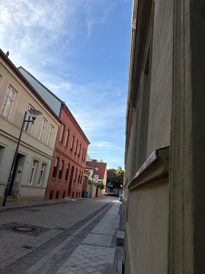 The image depicts a view of a street in Stendal, Germany, with buildings lining both sides. The street is paved with cobblestones, and there is a sidewalk on the left side. On the left side of the street, there is a light-colored building with several windows and a lamppost. Adjacent to it is a red brick building with arched windows. On the right side of the street, a beige building occupies the foreground, with its wall partially obscuring the view of the street and further buildings. The sky is blue with some wispy clouds. The overall scene appears to be a quiet, residential or commercial street in a European city.