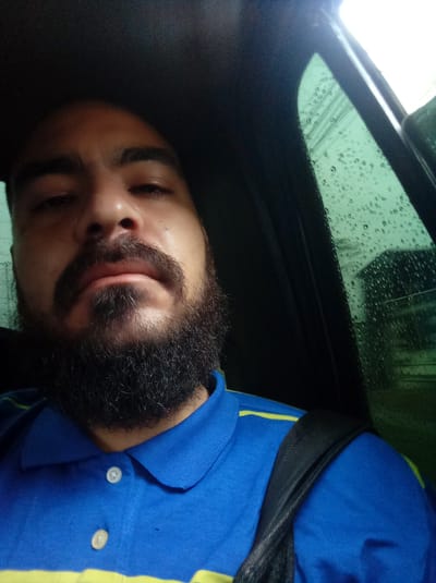 A bearded man wearing a blue and yellow polo shirt and a black shoulder strap is inside a vehicle. The car window is covered in raindrops, obscuring the view of a building outside. Based on the prompt specifying the location, this scene is in Parroquia Guatire, Venezuela.