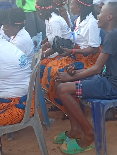 Several people are seated on plastic chairs in an outdoor setting. The individuals in the foreground are wearing matching white tops with a blue logo and vibrant orange patterned skirts. A woman is holding a black handbag. A young boy, dressed in a dark shirt and blue shorts, sits on a blue chair, wearing green sandals. The ground appears to be sandy or dirt. Some individuals are wearing red headbands.