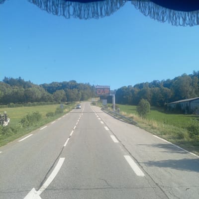 The image shows a view from a vehicle driving on a rural road in Laye, France. The road is paved with white dashed lines indicating lanes. On the right side of the road, there is a grass field and trees. Further ahead, there is a large electronic sign displaying "INFO ROUTE TOUR DE FRANCE 23/7 N85 FERMEE DE 13H A 17H CORPS-BRUT N". This indicates that Route N85 is closed due to the Tour de France on July 23rd from 1 PM to 5 PM. A white car is visible driving in the opposite direction on the road. The sky is clear and blue. There is a structure with a roof and fringe visible at the top of the image, suggesting it's the interior of a vehicle or a canopy.