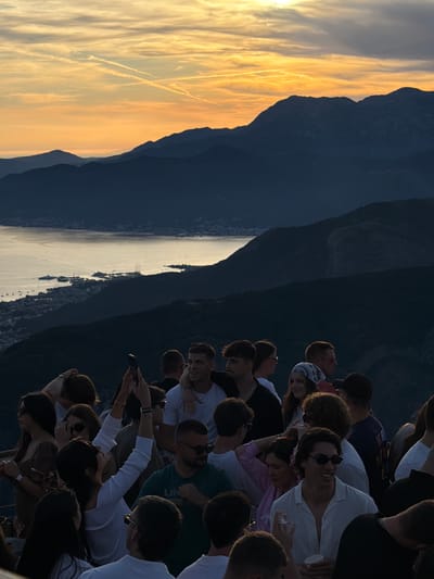 A group of people stand on a mountaintop overlooking a bay at sunset. The sky is filled with orange and yellow hues from the setting sun, casting a warm glow over the scene. Silhouetted mountains rise in the background, with a town and boats visible on the coast below. The people in the foreground are casually dressed, some holding phones and drinks. The overall atmosphere appears relaxed and social, with a sense of camaraderie amongst the group. The image captures a beautiful moment of natural beauty and human connection.
