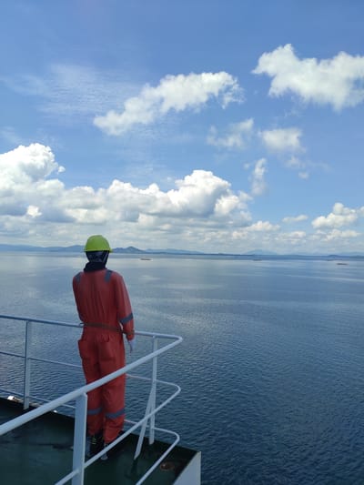 The image displays a mannequin dressed in safety gear on the deck of a ship, overlooking a vast expanse of water under a bright, partly cloudy sky. The mannequin, positioned upright near a white metal railing, wears an orange full-body suit with reflective grey stripes on the arms and legs, and a bright yellow hard hat. Its head is covered in black fabric beneath the hard hat. A rope appears to be loosely tied around the mannequin's midsection, securing it to the railing.

The setting is a calm body of water, likely the sea or a large bay, reflecting the clear blue sky and appearing smooth with gentle ripples. Along the distant horizon, a long stretch of land or mountains is visible, fading into the haze. Several small, indistinct objects, possibly other vessels, are dotted across the distant water. The sky is a vivid blue, filled with scattered, fluffy white cumulus clouds, indicating clear and sunny weather. The lighting suggests it is daytime, likely mid-morning or afternoon.

The scene evokes a maritime environment, possibly a safety drill or a display on a vessel sailing near Kunak, Malaysia. No visible text is present in the image.