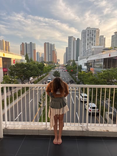 This is a vertical, wide-angle, eye-level shot of a young woman standing on a balcony, looking down at a street scene. The woman is positioned in the bottom right of the frame, with her back to the camera. She has long, brown hair and is wearing a casual outfit consisting of a dark denim skirt and a light-colored top. Her bare feet are visible.

The balcony she is standing on has a white metal railing and a concrete edge. Beyond the railing, a street unfolds with multiple lanes of traffic. Cars and a bus are visible on the road, and several pedestrians are walking on the sidewalk. Trees line the street, and a row of tall, modern apartment buildings forms the backdrop. The architecture of the buildings suggests an urban setting in South Korea, specifically Incheon, as indicated by the context.

The sky is partly cloudy, with a warm glow suggesting it is either early morning or late afternoon. The overall atmosphere is calm and observational. Some text is visible on the buildings in the background, including "PROGIO" on one of the apartment buildings and "SALE" on a sign below. The image captures a moment of quiet observation of urban life from a slightly elevated perspective.