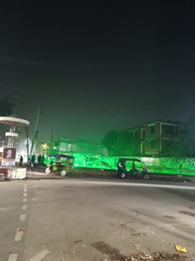 The image depicts a nighttime street scene in Lalmohan, Bangladesh. An asphalt road with faded white dashed lines occupies the foreground, with minor debris visible on the right shoulder. On the left, an elevated, light-colored circular structure is present, near which a banner displaying a woman's portrait is affixed to a pole. Several individuals, consistent with pedestrians, are congregated on the sidewalk area to the left. Two three-wheeled auto-rickshaws are on the road: one lighter-colored vehicle is positioned towards the center-left, and a darker auto-rickshaw is stationary or parked on the right side, with an individual standing near its rear. In the midground, a structure, possibly under construction or renovation, is prominently illuminated by a series of intense green lights, creating a horizontal band of green light across the scene. Scaffolding is visible around this green-lit structure. Behind it and to the right, a multi-story building with multiple window openings is partially visible, obscured by trees. The sky is dark, indicating nighttime conditions. Utility poles and overhead lines are also present.