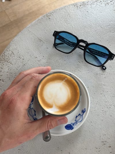 An overhead shot shows a hand holding a cup of coffee with latte art on a saucer with blue floral accents. The cup and saucer sit on a textured, light-colored surface next to a pair of black-framed sunglasses with blue lenses. The sunglasses are placed on the surface with the lenses facing upwards. Text on the sunglasses reads: "Mustella icon II AGGA BORN IN IBIZA" and "HANDCRAFTED IN ITALY 48024 CE CAT.2". The background shows a light-colored tiled floor. The image appears to be taken in Miami Beach, Florida, United States of America.