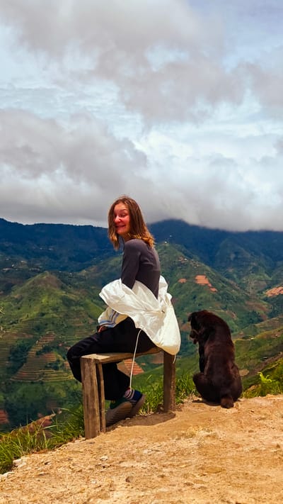 In Bắc Yên, Vietnam, a young woman with light brown hair, wearing a dark gray long-sleeved shirt, black pants, and a white jacket draped over her shoulders, sits on a wooden bench. She is looking back at the camera with a wide smile. Beside her, to her right, a dark brown dog sits facing away from the camera, looking out at the mountainous landscape. The environment consists of terraced hillsides covered in green vegetation and brown earth, indicating agricultural activity. The sky is filled with dramatic, overcast clouds. The foreground is a dirt path with scattered rocks and green grass. The scene is captured from a medium-wide shot, emphasizing the vastness of the natural scenery.
