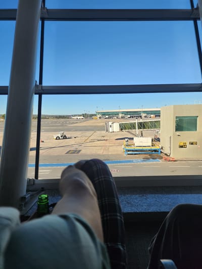 This media file shows an airport scene viewed from inside through a large window. The sky is clear and blue, suggesting a sunny day. The time of day appears to be daytime, likely late morning or early afternoon due to the strong sunlight casting shadows.
The scene depicts an airport tarmac with several elements visible. A jet bridge connects to an airport terminal building, which has several windows and signage. Various vehicles, including baggage carts and a pushback tractor, are present on the tarmac. In the distance, multiple aircraft can be seen parked at gates or on runways. The overall setting is an active airport.
In the foreground, parts of a person are visible. This includes their legs, one wearing plaid pants, and an arm holding a green glass bottle. The person appears to be relaxed, possibly waiting for a flight. The angle of the shot and the presence of the person's belongings suggest they are seated near the window.
The location is identified as Lago Sul, Brazil. There are some visible text elements, including numbers on the tarmac that appear to be gate or parking bay indicators, such as "14". There is also some text on the terminal building that is partially obscured and difficult to read clearly. The overall mood conveyed is one of waiting and observation within an airport environment.