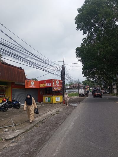 A street scene under a cloudy sky. Two women, wearing traditional clothing, walk along the sidewalk near a red and yellow food stall labeled "BAKSO SOLO". Several motorcycles are parked nearby. An asphalt road runs beside the sidewalk, with various vehicles driving on it. Overhead, a dense network of electrical wires creates a chaotic web against the sky. A large tree stands to the right of the road.