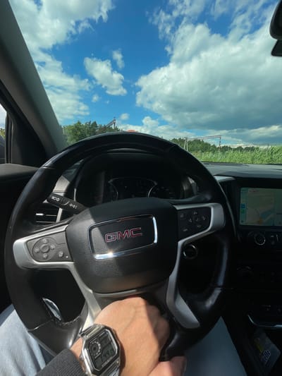 This media file captures a first-person perspective from inside a GMC vehicle. The main subject is the steering wheel with its prominent GMC logo and control buttons. A person's hand, wearing a Casio digital watch, is resting on the steering wheel. The watch displays the time as 1:44:19, with "13:43" also visible, along with "LTE" and a map on the car's infotainment system.

The setting is a roadside view through the windshield, indicating the vehicle is in motion. The scene outside features a bright, cloudy sky with patches of blue, suggesting it's daytime with fair weather. Green fields and trees are visible along the side of the road, with a power line structure in the distance.

The overall mood is casual, possibly indicating a road trip or daily commute. The rotation of the image suggests it was taken while the vehicle was turning or was oriented at an angle. The visible text includes brand names like GMC and Casio, along with standard car interface text and numerical data. The presence of "Voskresensk, Russia" as location context provides geographical information for the scene.