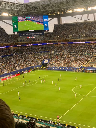 This is a high-angle, wide shot of a soccer game at Hard Rock Stadium in Miami Gardens, Florida, USA. The stadium is filled with spectators, most of whom are wearing light blue clothing, suggesting they are supporters of one of the teams. A large video screen at the top of the stadium shows the game's score, with Saudi Arabia and Uruguay tied at 1-1. The field is green with white lines, and several players in white and dark uniforms are visible. In the foreground, a crowd of spectators is gathered, with some wearing hats. The lighting indicates it is late afternoon or early evening. The overall scene depicts a professional soccer match being played in a large, modern stadium.