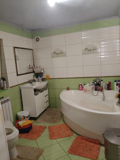 The image depicts a bathroom in a residential dwelling in Chornobaivka, Ukraine. The bathroom features light green tiled walls, with the lower half a solid color and the upper half featuring a floral pattern. A white corner bathtub with a silver faucet is prominent. To the left of the bathtub, there is a white sink with a silver faucet, mounted on a white vanity with drawers. A mirror with a dark frame hangs above the sink, reflecting the contents of the vanity. To the left of the sink, a white radiator is visible. A white toilet is situated in the lower left corner of the image, partially obscured by a doorway. The floor is tiled in a light green. Several orange bath mats are placed on the floor, one near the toilet and two near the bathtub. A large, silver trash can with a lid stands to the right of the bathtub. Various toiletries and cleaning supplies are visible on the shelves and around the sink and bathtub. The lighting in the room appears to be from an overhead fixture, casting a bright illumination. There are no people visible in the image.