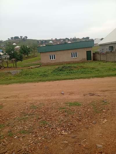 A wide shot captures a slightly hazy, overcast day in a rural area, likely Ashata, Nigeria. The foreground is a dusty, brown dirt road scattered with small stones and patches of sparse green grass. To the right of the road, a low, rough brick wall borders a yard. Beyond this wall, a light grey building with a white, corrugated metal roof stands prominently. This building has two rectangular windows with white frames and a single dark green door with a decorative pattern. Adjacent to this, on the left side of the frame, is a smaller, tan-colored building with a slanted, dark green metal roof. This building also features two rectangular windows with white frames. Behind these structures, the landscape slopes upward, revealing more houses with similar metal roofs nestled amongst trees and rocky terrain. The overall impression is of a developing residential area on a hillside under a muted sky.