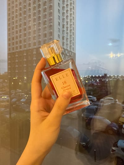 A hand holds a rectangular glass perfume bottle with a gold cap and a light peach label. The label has black text that reads "ELLE 18 Cherry desire". The bottle contains a red liquid. In the background, a tall, modern building with many windows is visible. This building is reflected in a large glass window, which also shows a cityscape at dusk with lights from cars and buildings. The location is identified as Drozhzhino, Russia.