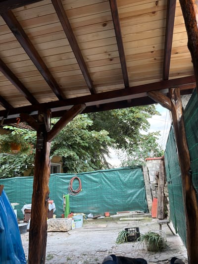 The image depicts an outdoor scene, likely a backyard or a workshop area. The foreground is dominated by the rustic wooden structure of a roof or pergola, with dark wooden beams supporting a ceiling of light-colored wooden planks. These beams have a natural, unfinished appearance, some showing rough textures and knots.
Beyond the wooden structure, the scene opens up to a more cluttered outdoor space. A large area is covered by a green tarp, suggesting a construction or storage area. Various objects are visible in front of and around the tarp, including gardening tools, containers, and what appear to be potted plants. There's a coiled orange hose hanging on the tarp. In the background, lush green trees and foliage are visible, with a partly cloudy sky overhead. The ground is paved with stones.
There are no people clearly visible in the image. The overall impression is one of a functional, somewhat unkempt outdoor area. The lighting suggests it is daytime, possibly late morning or afternoon, given the shadows and the brightness of the sky. The presence of greenery and the open-air setting point to a location in a rural or semi-rural environment.
No visible text is present in the image.