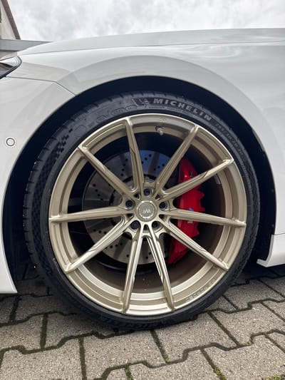 This is a close-up, vertically oriented image of a car's wheel and tire, set against a backdrop of a cobblestone driveway. The car is white and appears to be a high-performance vehicle, evident from the visible red brake calipers behind the intricate, multi-spoke alloy wheel. The wheel itself is a light gold or bronze color.
The tire is a Michelin Pilot Sport 4S, with detailed white lettering visible. Text on the tire includes "MICHELIN," "285/30 ZR 20 (99Y) EXTRA LOAD," "OUTSIDE," "TREADWEAR 300," "TRACTION AA," "TEMPERATURE A," and "DOT 16U UK 025X 0626." The rim of the wheel also has embossed text reading "RS FORGED." A small "WE" logo is visible on the wheel spokes.
The setting suggests an outdoor location, possibly a residential driveway or a parking area, given the cobblestone pattern. The lighting indicates it is daytime, and the sky, visible in the upper left corner, is overcast, suggesting cloudy weather. There are no people in the image. The focus is entirely on the automotive detail, highlighting the wheel, tire, and braking system. The overall impression is one of automotive craftsmanship and performance.