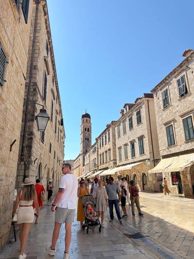In Dubrovnik, Croatia, a bright, sunny day illuminates a bustling pedestrian street lined with stone buildings. The architecture features large, rough-hewn stone blocks and dark-shuttered windows, characteristic of the Old Town. In the distance, a prominent bell tower with a dark dome rises above the buildings, identifying the location as Stradun, the main street of Dubrovnik. A diverse group of tourists are walking along the street. Some are dressed in casual summer attire, while others are pushing a stroller with a baby. Several shops with awnings are visible along the right side of the street. The paved ground is made of large, irregular stones. The scene captures the vibrant atmosphere of a popular tourist destination.