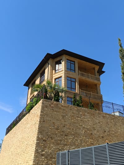The image displays a modern, multi-story building constructed with light-colored stone, situated on a raised stone foundation. The building features multiple balconies with ornate railings and large windows with dark frames. It is surrounded by greenery, including palm trees and shrubs, and protected by a black metal fence. A tall, slender evergreen tree stands to the right of the building. The scene is set against a clear, bright blue sky. The building's architecture suggests it is a residential property, likely a villa or a luxurious home, in a warm climate. Based on the location provided, this is a property in Verkhnevesyoloye, Russia.