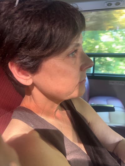 A medium shot, side profile of a woman with short, dark hair is captured from the chest up. She is seated indoors, with a dark red upholstered seat visible on the left. Her skin appears to be sun-kissed, with a golden hue and some faint red undertones. She is looking off to the right, her gaze directed towards a window.

Through the window, a blurred background of green foliage suggests an outdoor environment, possibly trees or bushes, bathed in bright sunlight. Inside the vehicle, a dark surface, possibly a seat or armrest, is visible in the background to the right. The lighting suggests it is daytime, with sunlight streaming in, creating shadows on the woman's face and shoulder. The overall impression is that of someone traveling by vehicle, perhaps a bus or a car, on a sunny day.