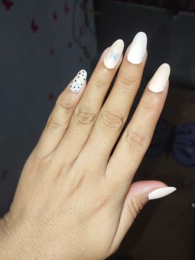 A person's hand with manicured fingernails is shown. The nails are an almond shape and painted in various colors and designs. The index finger has a white nail with a light blue butterfly decal. The middle finger is painted white. The ring finger has a light beige nail with blue polka dots. The pinky finger has a light beige nail. The thumb also has a light beige nail. The background is dark and out of focus, with some blurred shapes that might be furniture or fabric. The image appears to have been taken indoors. There is no information available about the location in Punta de Piedras, Venezuela.