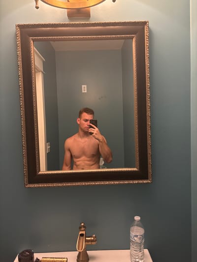 A young man with short, curly brown hair and a muscular physique is taking a mirror selfie in a bathroom. He is shirtless and wearing what appears to be patterned boxer briefs. He is holding a black smartphone in his right hand, angled to capture his reflection. A thin gold chain is visible around his neck. The bathroom features a dark, ornate framed mirror above a vanity with a sink. The walls are painted a teal blue. A brass faucet is visible on the vanity, along with a dark jar and a water bottle. In the reflection, an electrical outlet and a light fixture are visible on the wall behind him.