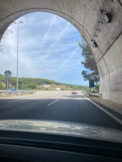 The image depicts a forward-facing view from inside a vehicle exiting a concrete archway structure, likely a tunnel or underpass, onto the C-32 highway near Sant Pol de Mar, Spain. The archway's interior is concrete, displaying seams and attached metal grating with two overhead lighting fixtures on the right side.

The open highway ahead is asphalt, featuring at least two visible lanes of travel marked by white solid and dashed lines. A light-colored compact car, possibly beige or pink, is observed in the distance in the left lane, moving away from the viewer. Further to the left, a dark blue vehicle is partially visible behind a large, circular grey road sign, which appears to be a regulatory sign with an obscured symbol.

Along the left side of the highway, multiple tall grey streetlights with dual luminaires are spaced intermittently. The terrain beyond the left shoulder comprises a vegetated hillside, densely covered with green trees and shrubs, and some lighter patches of dry vegetation or exposed earth. A concrete barrier separates the highway from the embankment.

On the right side, immediately outside the archway, a large evergreen conifer tree with dense foliage is prominent. Beneath it, a blue rectangular road sign indicates "C-32" and "km 110", confirming the location on the C-32 highway in the Sant Pol de Mar area of Catalonia. A metal guardrail runs along the right shoulder.

The sky is light blue with scattered, thin white clouds, suggesting clear weather. A faint, linear contrail is visible in the upper left sky. The lower foreground of the image shows the dark dashboard and windscreen reflection of the viewer's vehicle. No people are discernible.