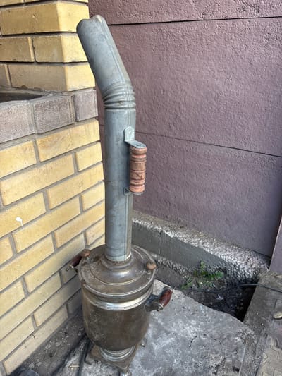 A weathered, metal samovar with wooden handles stands on a concrete surface next to a yellow brick wall. A grey metal pipe, likely a chimney or vent, extends upwards from the top of the samovar and bends to the left. The pipe is secured to the samovar by a metal bracket with a wooden handle. The background consists of a textured, dark brown wall. The ground around the samovar is a mix of dirt, small stones, and some concrete debris. There are no people visible in the image. The setting appears to be an outdoor urban environment, possibly an alleyway or backyard in Chernogorsk, Russia.