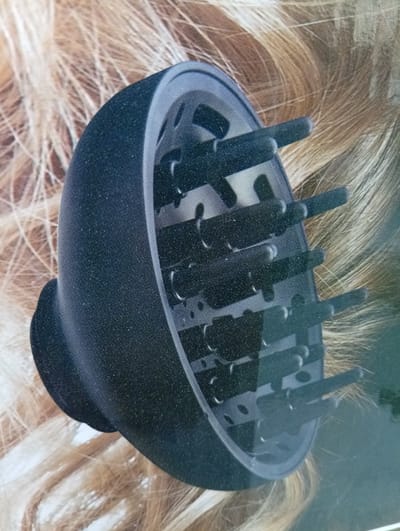 A black hair dryer diffuser attachment is positioned on a background of blonde hair. The diffuser has a rounded shape with numerous prongs extending inward. The lighting is bright, casting a slight shadow on the left side of the diffuser. The image appears to be a product shot, likely for advertising purposes, and does not depict any specific location in Rezina, Moldova or any people.