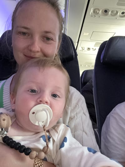 The image shows a woman holding a baby in her arms. The woman is seated on a plane. She is wearing a cream-colored sweater, and the baby is wearing a white onesie with blue polka dots. The baby is sucking on a white pacifier. The woman is smiling, and the baby is looking at the camera. The image appears to be a selfie taken on a plane. The overhead compartments are visible, and the seats are blue. The woman has blonde hair, and the baby has light brown hair. The woman is wearing a gold bracelet on her left wrist. The baby has a black and wood beaded chain attached to its clothing. The coordinates 31.998, 34.874 correspond to a location near Ben Gurion Airport in Israel. It is likely that the image was taken on a flight that was departing from or arriving at this airport.