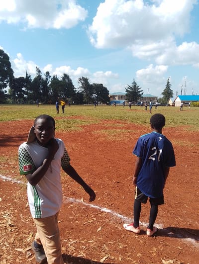 Two young men stand on a dirt field in Rungiri, Kenya. The man on the left is wearing a white and green shirt, beige pants, and black shoes. He has his hand on his chin and is looking at the camera. The man on the right is wearing a blue jersey with the number 21 on the back, black pants, and white sneakers. He is looking towards the left.  The field is red dirt and has a white line on the ground. There are several people playing soccer in the background. The sky is blue with white clouds.  There are trees and buildings in the distance.