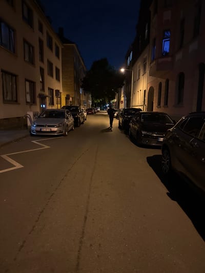 A person walks down a residential street at night in Duisburg, Germany. The street is lined with apartment buildings on both sides, with cars parked along the curb. The sky is dark blue, indicating it is nighttime. The street is illuminated by streetlights, casting long shadows. The person is walking away from the camera and appears to be alone. The overall scene is quiet and still, with no other people or activity visible.