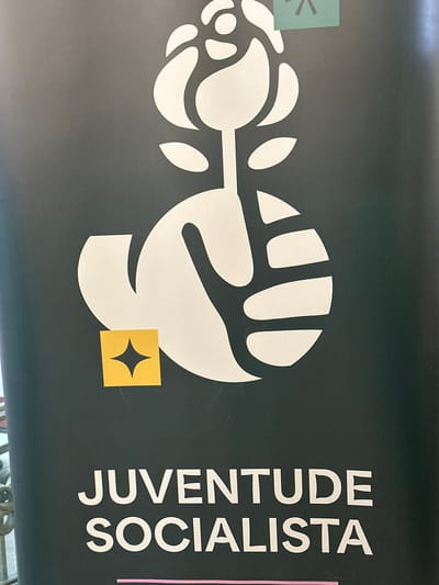 The image displays a vertical dark-colored banner, likely dark green, featuring white graphics and text. The upper portion of the banner presents a stylized white logo depicting a rose atop a stem, which is grasped at its base by a white hand. Below this logo, the words "JUVENTUDE SOCIALISTA" are printed in white uppercase letters. A small light-green rectangular sticker with a white asterisk-like symbol is affixed to the banner's upper right corner. Additionally, a small yellow square sticker with a black four-pointed star is placed over the lower-left section of the white hand logo. Minor surface imperfections and creases are visible on the banner. A portion of a dark, ornate metal structure, possibly a railing, is visible along the bottom left edge of the frame. The banner is located within the city of Aveiro, Portugal.