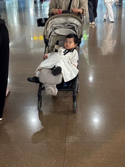 A young child is sitting in a stroller in an airport terminal. The child is wearing a white outfit and black shoes. The stroller is gray and black. In the background, a person is pushing the stroller, and other people are walking in the distance. The floor of the airport terminal is tiled and reflective. There are signs with Korean writing on the wall. The location is the Incheon International Airport in Incheon, South Korea.