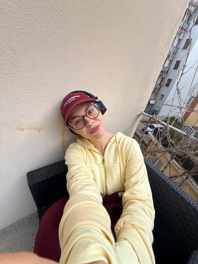 The image features a person, likely a young woman, captured in a selfie-style shot in an outdoor setting in Sanremo, Italy. The photograph is taken from a low angle, with the subject positioned on their side due to the image's rotation.

The person is wearing a light yellow, long-sleeved zip-up hoodie, round-framed glasses, and black over-ear headphones. They also have on a maroon baseball cap with white embroidered text that reads "JAMSBURG WILLIAMSBURG BROOKLYN". Their expression is neutral, with a slight hint of a smile, as they look directly into the camera.

They appear to be seated on a dark, possibly black, woven outdoor chair or furniture, a part of which is visible in the lower right quadrant of the frame. A light beige, textured wall occupies the left side and foreground of the image, showing some slight discolored marks.

In the background, above the wall, there are numerous tangled, bare branches, suggesting a vine or dormant tree. Beyond these branches, other buildings with windows and balconies are visible under what appears to be a bright, possibly overcast, daytime sky. The setting suggests a balcony or terrace overlooking an urban residential area.